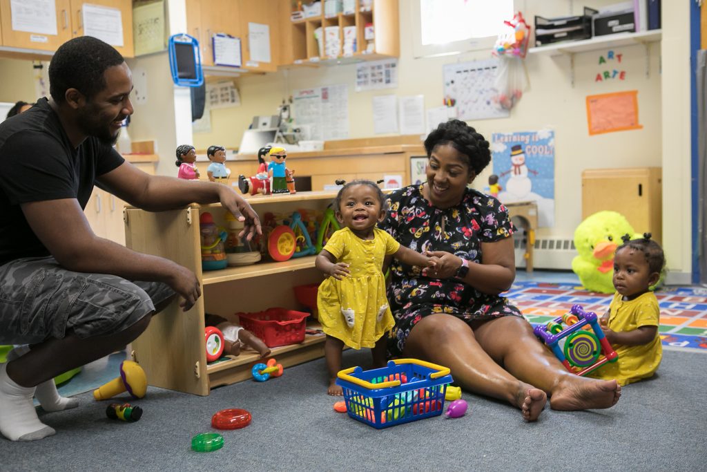 Mother sits on the floor of a child care center with her twin daughters while their father smiles