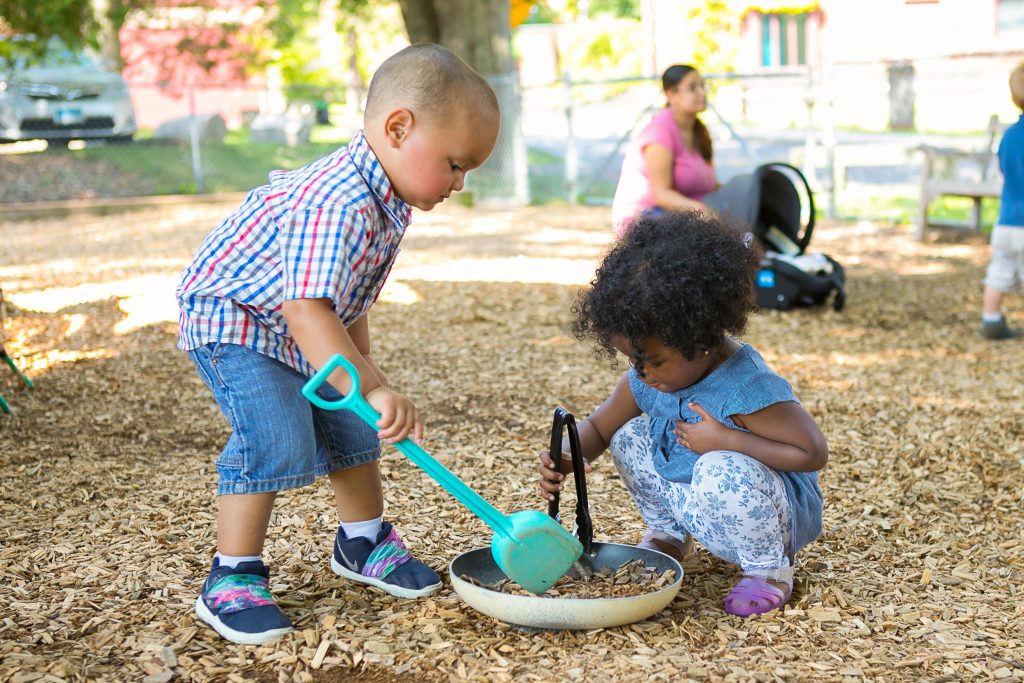 Two toddlers play with plastic shovels on a playground