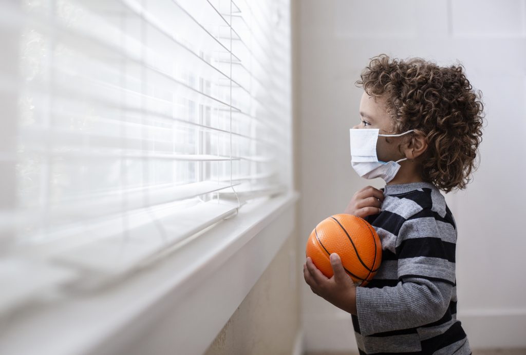 Child wearing a mask looks outside longingly.