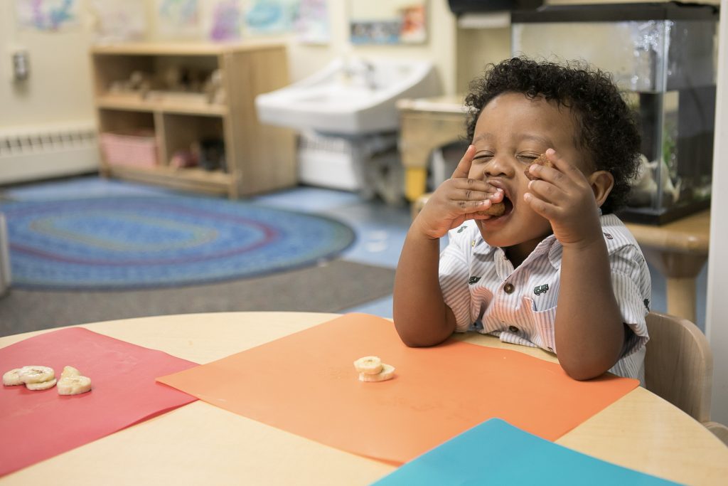 Toddler sits at a table with his eyes closed, joyously eating banana slices